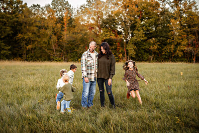 Family of six in a grassy field. The children are running circles around the mom and dad.