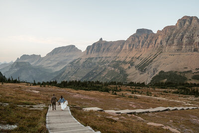 A couple walks hand in hand along a wooden boardwalk surrounded by sweeping mountain views and golden light during their adventurous Glacier National Park elopement, captured by Sydney Breann Photography.