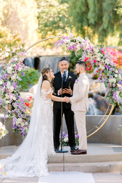 A couple holds hands during a wedding ceremony under a floral arch with pink, purple, and white flowers, set outdoors near a waterfall.