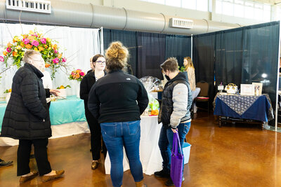 two people standing at a vendor table with flowers behind the table area