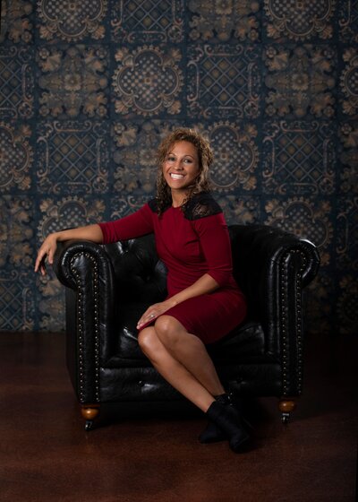 Personal fitness trainer Jackie Collins sitting on a black chair with an embellished black backdrop smiling wearing a red and black knee length dress.