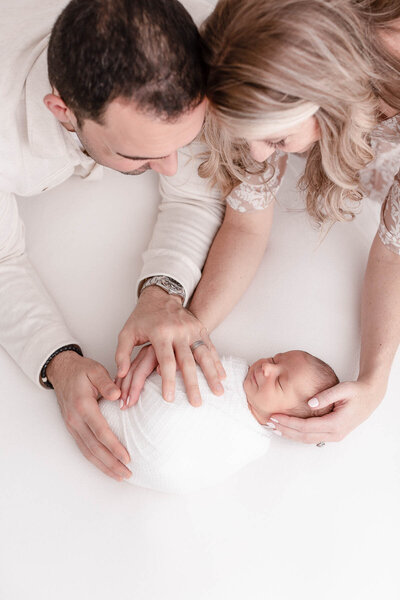newborn baby wrapped in white on a cream backdrop with parents looking down at him and both parents have their hands on him. Parents are dressed in light, neutral tones.