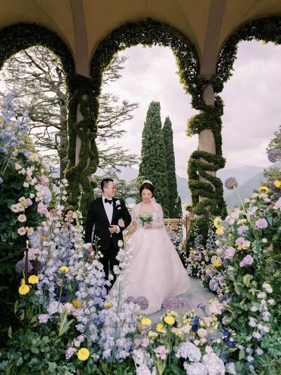 A bride in a wedding dress stands in a grand doorway in villa Sola Cabiati, gazing towards a scenic view of mountains and Lake Como. The elegant room features ornate decor, vases, and cushioned benches, capturing the essence of an Italy Wedding Photographer amid the serene outdoor backdrop.