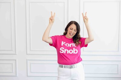 Brand designer in hot pink Font Snob t-shirt celebrating with playful peace signs against white wall during brand photoshoot