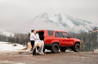 A bride and groom sit on the tailgate of their vintage red Toyota on their mountain wedding day in the winter. 