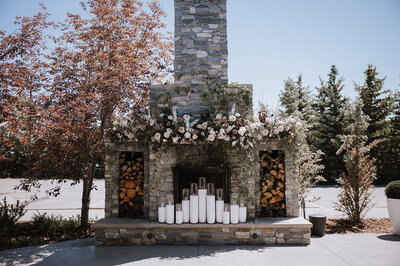 Outdoor ceremony set up in front of fireplace with lush florals and large candles on the mantle at Flores and Pine in Calgary