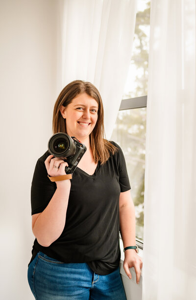 Ashley Morrisey standing next to a window with sheer curtains holding her camera