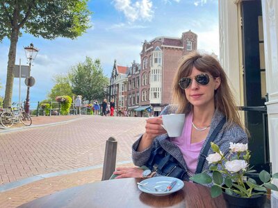Award-winning luxury travel expert Stephanie Dosch enjoying coffee at a canal-side café in Amsterdam, Netherlands, with traditional Dutch townhouses and a brick-paved street in the background ©Stephanie Dosch | theViatrix Luxury Travel