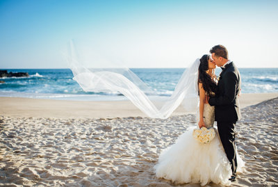 Newly married couple share a romantic kiss on the beach as her veil flies in the wind