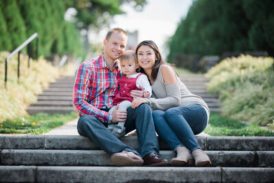 Heartwarming Family Photoshoot on Park Steps: Husband and wife gracefully seated, tenderly cradling their daughter, capturing a moment of pure love