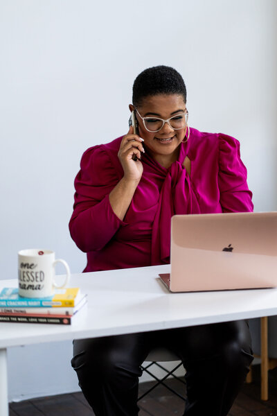 Career strategy coach calling an important client while working in her professional office. She sits at a white desk wearing an elegant pink outfit, using her laptop and phone as essential job tools.