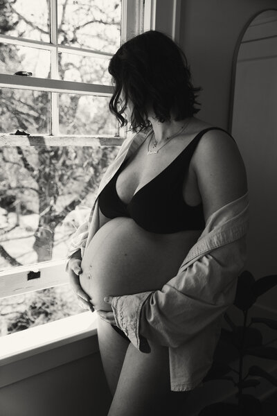 Black and white image of pregnant woman in underwear looking out the window