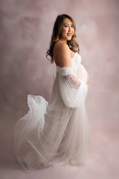 Pregnant mom sitting on a posing block gently touching her bump while looking off to the side.