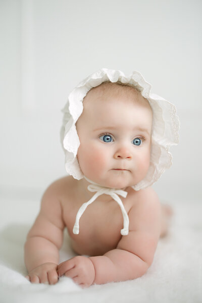 an 8 month old baby lays on her tummy in a white studio while wearing a white baby bonnet. Her blue eyes shine in the photo and the smirk on her face is adorable.