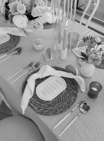 A close-up of a bohemian guest table at a National Arts Centre wedding, featuring beige textured linens with a series of taupe tapered candles surrounded by white bud vases with peach, cinnamon and white fresh flower booms and a modern half-circle table number. Each place setting has a knotted white linen napkin and an oval menu on top of a woven rattan charger plate with gold and white cutlery on each side.