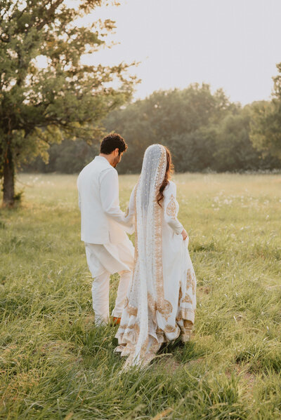 Couple in traditional South Asian wedding attire walking hand in hand through a sunlit field, with the bride wearing an embroidered veil and gown.