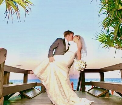 groom holding bride and kissing on beach dock under palm trees