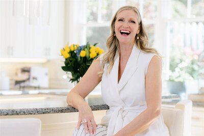 Phillipa Leseberg in a white outfit, an accomplished author, sits on a chair in a bright kitchen adorned with yellow flowers on the counter behind her.