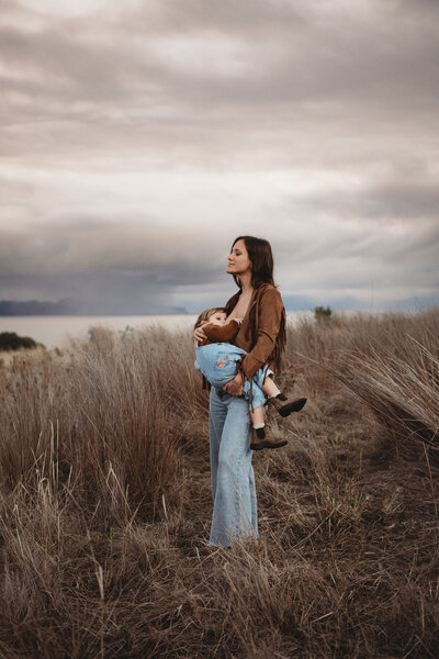 A woman in a brown jacket and jeans holding a child, standing in a grassy field under a cloudy sky, perfectly capturing the essence of South Coast motherhood portraits.