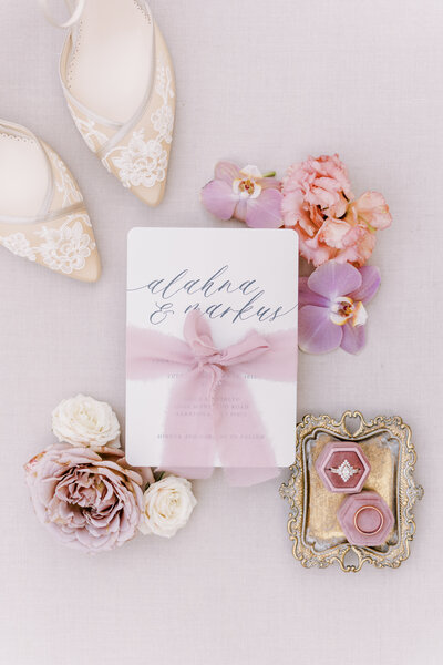 a wedding invitation in the middle of florals, the brides bella belle wedding shoes, and blush velvet ring box.