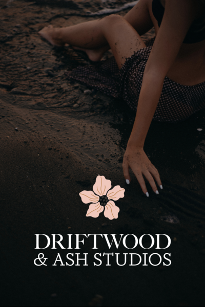 Dramatic dark photo of a woman cropped from neck down on the beach with a pink flower and the words Driftwood & Ash Studios