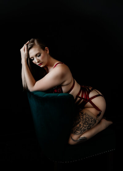 Blonde woman in red strappy lingerie kneeling on a green chair with a black background. Her elbows are resting on the back of the chair and her hands are in her hair. Her eyes are closed giving the photo a dark and moody feeling.