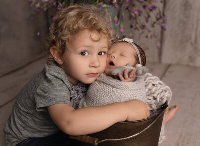 toddler holding his sister that is wrapped in purple knit fabric