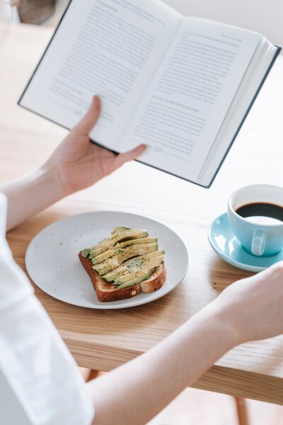 Self care image of woman's hand holding book with plate of avocado toast and cup of coffee