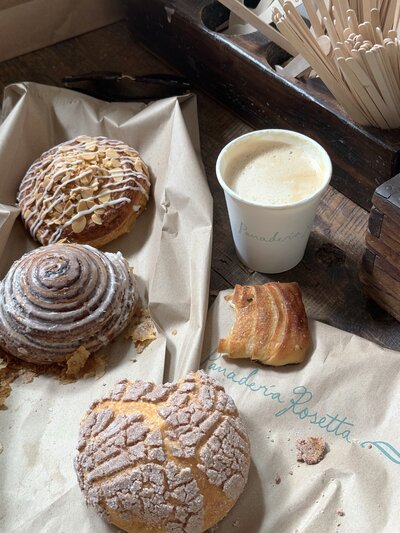 A selection of handcrafted pastries from Holly’s Bakery displayed on branded bakery paper. A steaming cup of coffee sits beside the assortment, with wooden stirrers and napkins nearby. A visually appealing presentation that highlights the bakery’s high-quality offerings and artisanal touch.