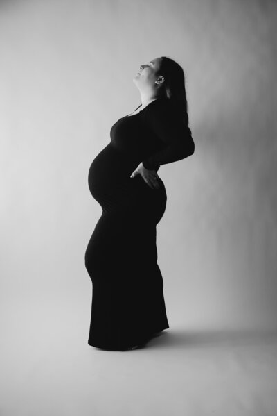 Black and white glamour maternity photo of mom in black dress and standing in a studio in Maryland.