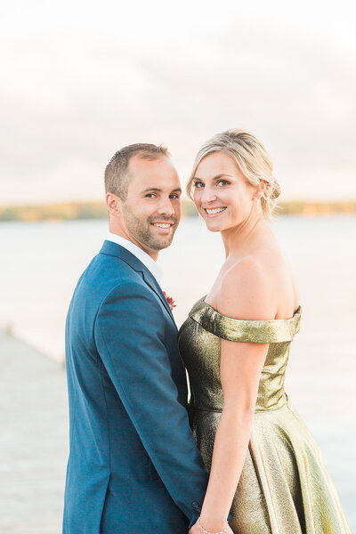 Bride in gold wedding derss next to groom in navy suit on the dock of a lake.