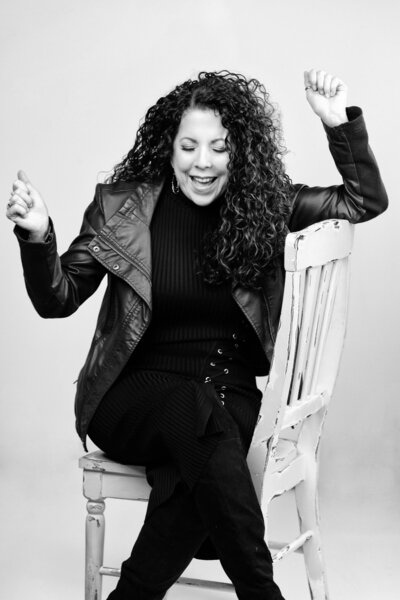 Black and white image of a woman with curly hair sitting on a chair with her eyes closed, smiling and raising her hands joyfully. She is wearing a leather jacket. A text overlay at the bottom reads, "SUBSCRIBE ON YOUTUBE.
