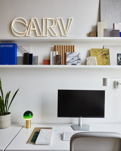 A white chair and desk with a computer monitor and green plant accents positioned against a wall with floating shelves containing various books, art pieces, and CARV's logo.