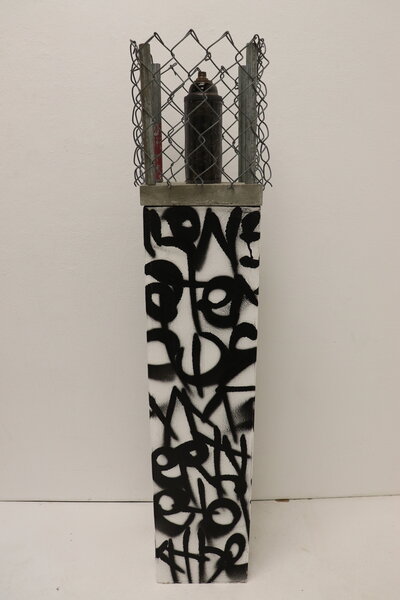 A dark ceramic spray paint can is decorated with a carving of young children and sits atop a graffiti painted pedastal, but is caged in by a wire fence