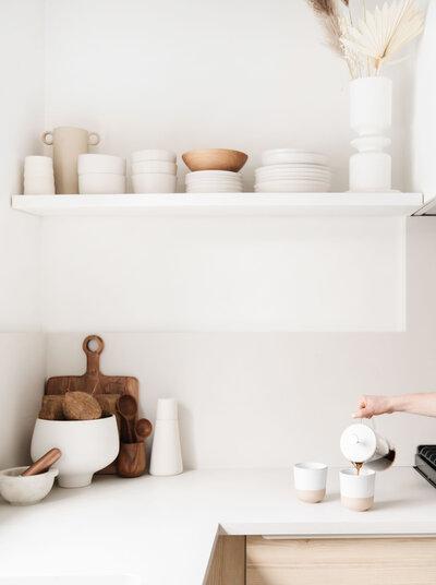 white kitchen counter, white walls, white floating shelf with neutral clay dishware, white bowl with coconuts, wood cutting board, white mugs with hand pouring coffee from french press