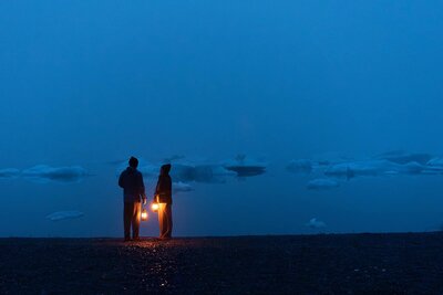 It is night time and the couple who just had a helicopter adventure elopement that day is walking next to Bear Lake in the dark, exploring from the glamping yurt included in their elopement package.  They are each carrying a lantern that glows with warm light in the dark blue of the night.  They are silhouetted against the foggy background of the lake, where shadows of icebergs and are visible all across the water.  
