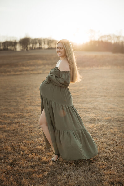 Pregnant woman in flowing green dress stands in open field duirng golden hour in Bucks County PA 