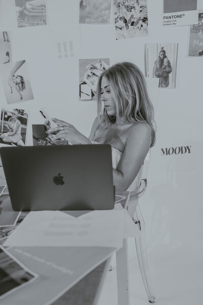 ALT: Grace sitting at a table looking at her phone with her laptop up and client papers around her