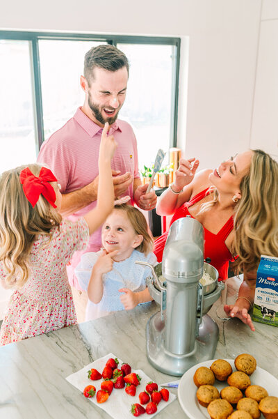 Family of four, pregnant mother in red dress, dad and two daughters baking cupcakes in a white modern kitchen