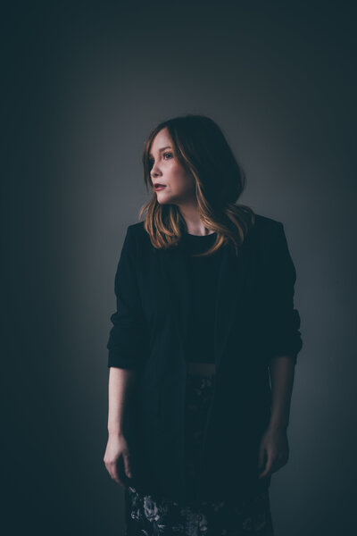 A woman with brown hair wearing a black blazer stands against a dark, neutral background, looking off to the side, exemplifying the professionalism of an online business manager.
