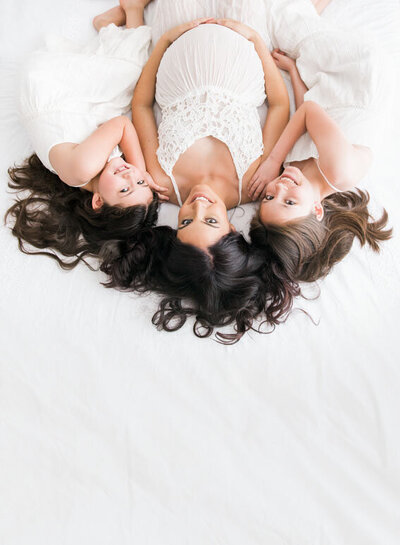 Joyful maternity portrait of a mother in a lace gown lying on a bed with her two daughters, captured in natural light by a Destin maternity photographer.
