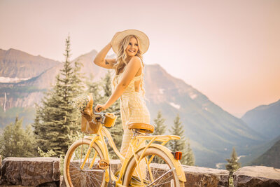 senior girl with long blonde hair and a cream hat holding on to a yellow bike at glacier national park