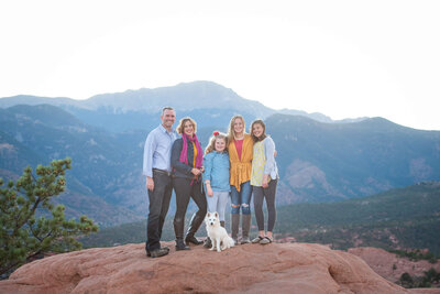 A family of 5 stand on a red rock with their small white dog