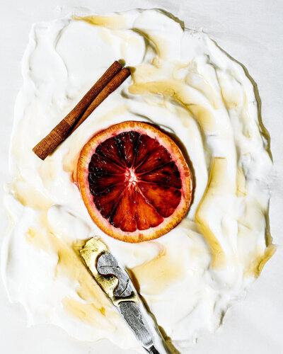 A vibrant and artistic overhead shot of creamy yogurt topped with a blood orange slice, a cinnamon stick, and a drizzle of honey. Captured by Mica McCook, this image highlights the contrast between the bright, fresh ingredients and the smooth yogurt base.