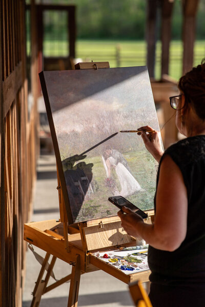 Artist painting a wedding scene on a canvas outdoors, using a photo for reference