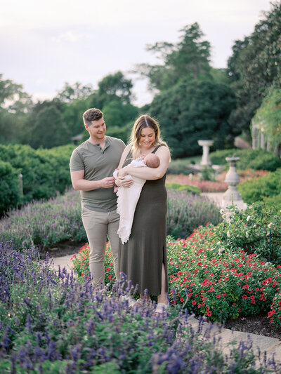 A mother standing in a garden holding her newborn as the mother and father both look at the newborn by Katie Stansfield Photography, a Richmond newborn photographer.