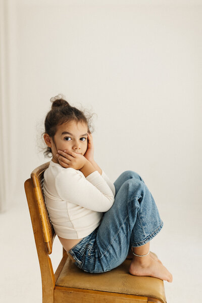 A little girl wearing 90s styled jeans sitting on a chair during a studio family photoshoot in christchurch