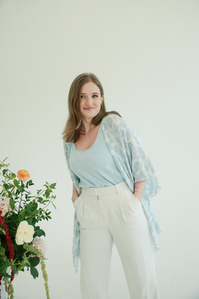 Dania, a wedding content creator with long hair sits smiling on a green armchair, wearing a cream top and rust skirt, surrounded by soft pink and white flowers indoors.