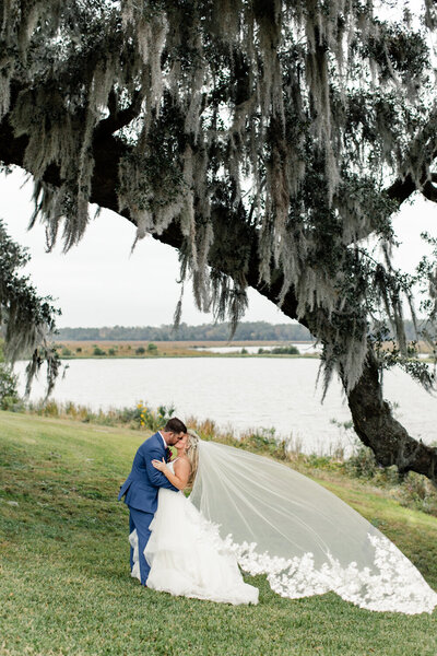 Bride in white gown and Groom in blue suit kissing under an oak tree at the Middleton Place Charleston Venue captured by Charlotte wedding photographer.