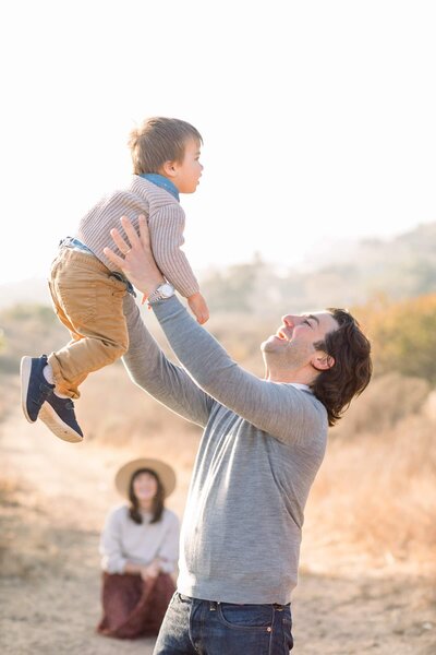 A father holds his son up in the air as the mother watches from the background